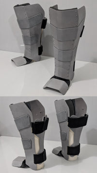 A pair of gray metal protective shin guards with black straps, shown from multiple angles on a white surface against a light gray background.