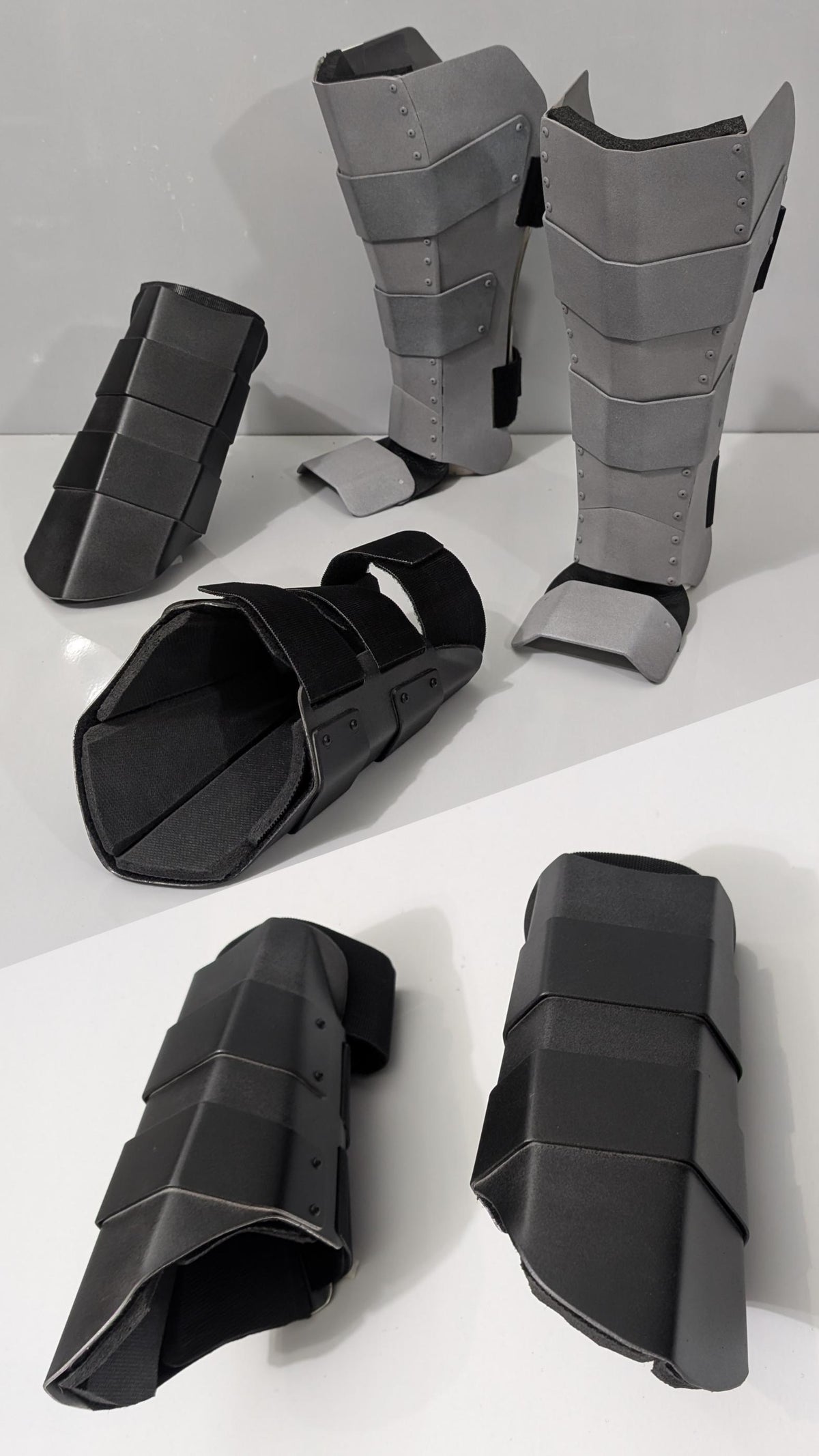 A set of black and gray padded metal armor or shin/ankle guards arranged on a white surface against a white background.