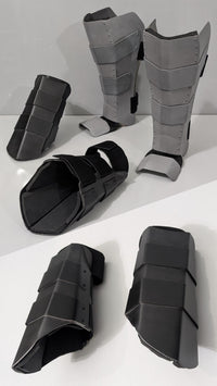 A set of black and gray padded metal armor or shin/ankle guards arranged on a white surface against a white background.