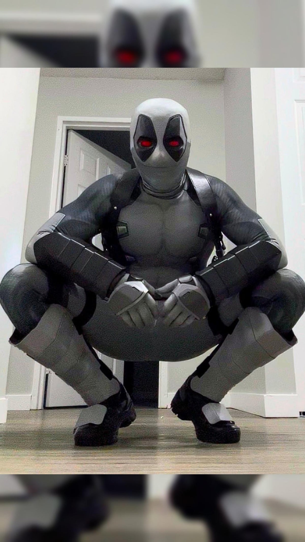 A person wearing a full Deadpool costume crouching indoors in front of white doors. The costume includes metal shin guards and full protective metal gauntlets