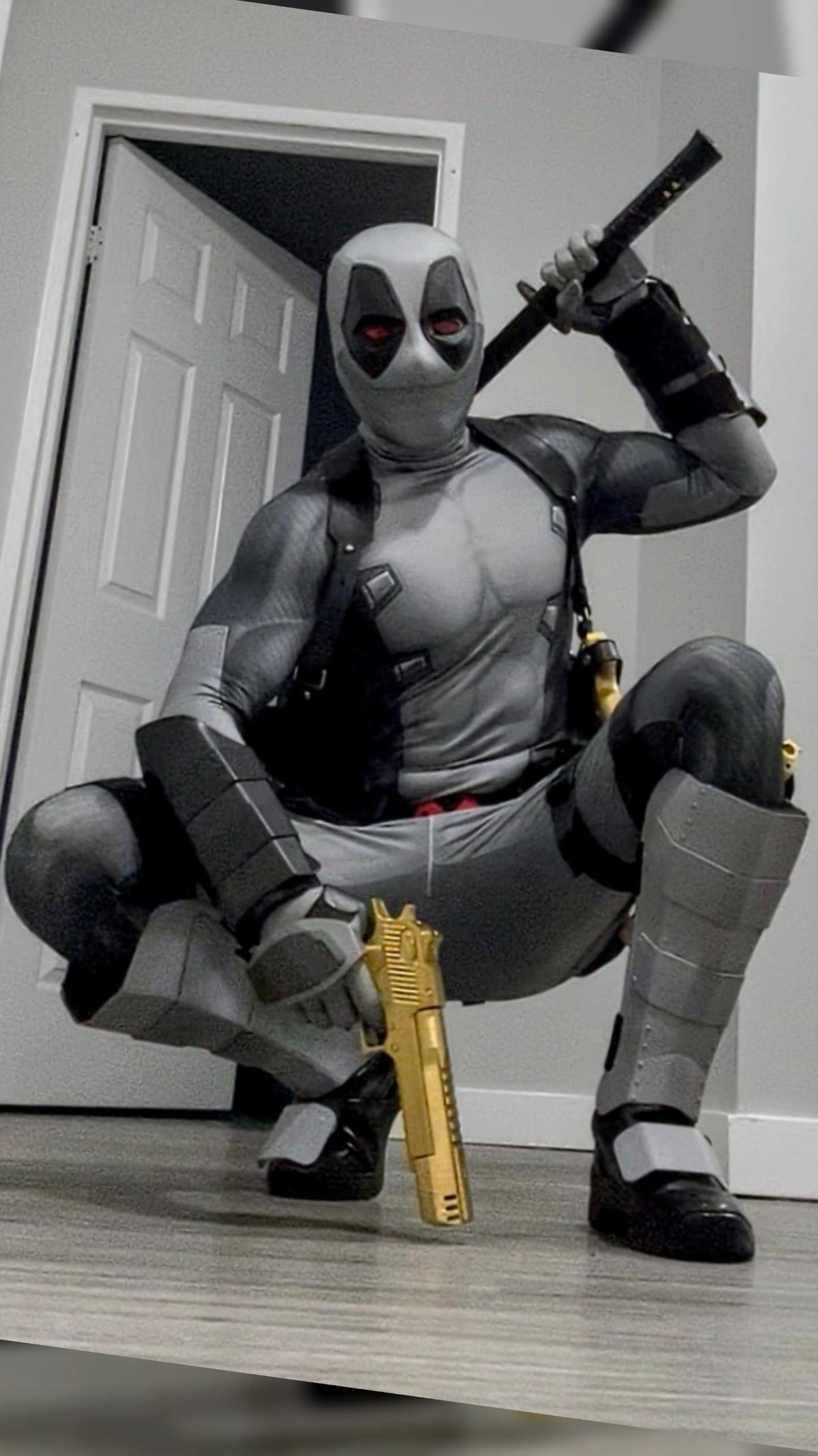 A person crouching wearing a full Deadpool costume indoors in front of white doors. The costume includes metal shin guards and full protective metal gauntlets
