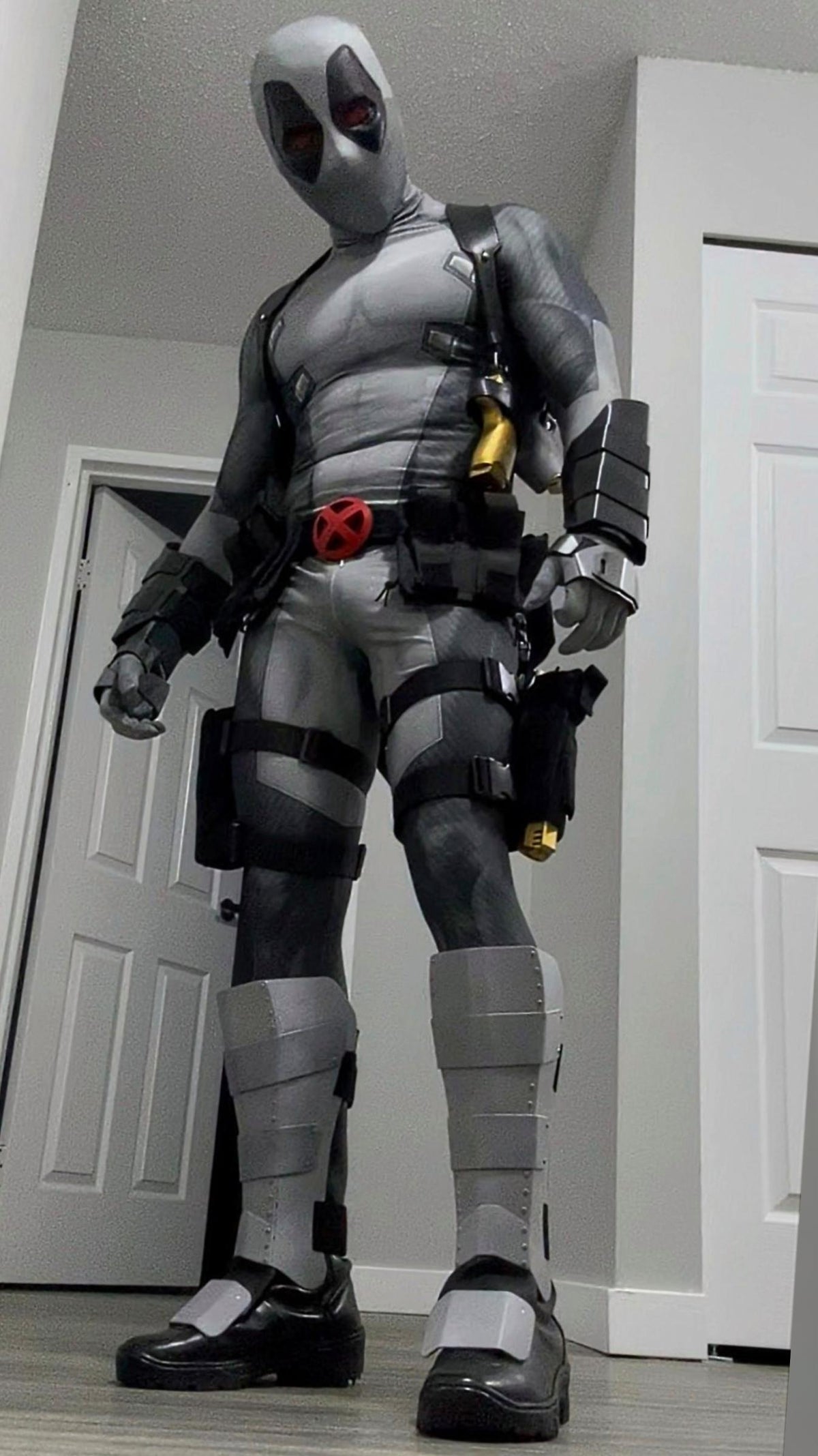 A person wearing a full Deadpool costume standing indoors in front of white doors. The costume includes metal shin guards and full protective metal gauntlets